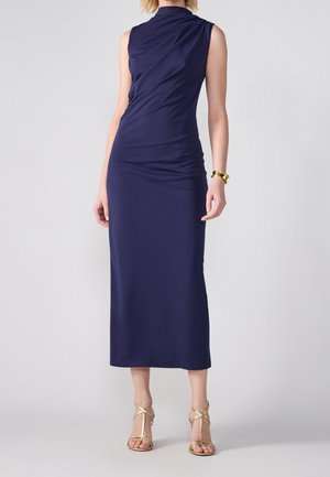 Kobieta w granatowej sukience midi bez rękawów z drapowanym dekoltem i złotych, paskowych sandałach na wysokim obcasie, stojąca z jedną ręką w luźnej pozycji.