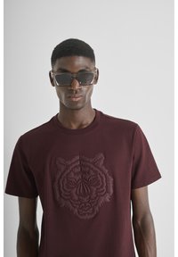 Bordo pamučna majica s reljefnim uzorkom tigra na sredini, okrugli izrez i kratke rukave. Model nosi tamne sunčane naočale.