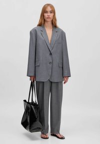 Grauer übergroßer Blazer mit zwei Knöpfen und Fronttaschen, kombiniert mit passenden grauen Hosen und einer glänzenden schwarzen Tote-Bag. Flache Sandalen.