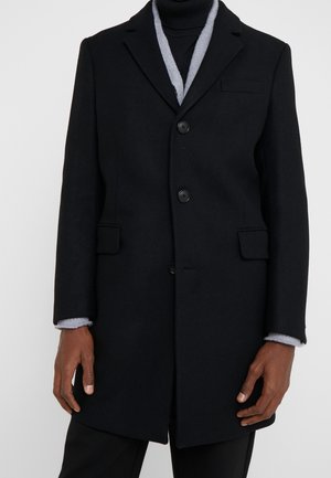 Czarny zapinany na guziki płaszcz z klapami noszony na szarym swetrze i czarnym golfie, model stoi z rękami swobodnie opuszczonymi wzdłuż ciała.