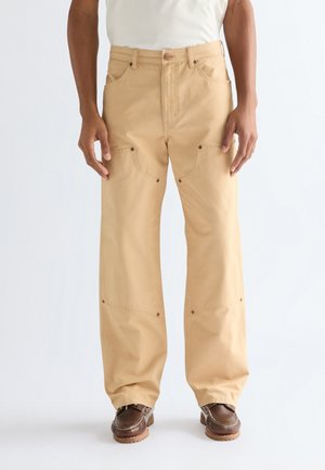 Mies, jolla on beigejä leveälahkeisia housuja, joissa on vahvistetut polvipaikat, ja ruskeat nahkakengät, seisoo yksinkertaisen valkoisen taustan edessä.