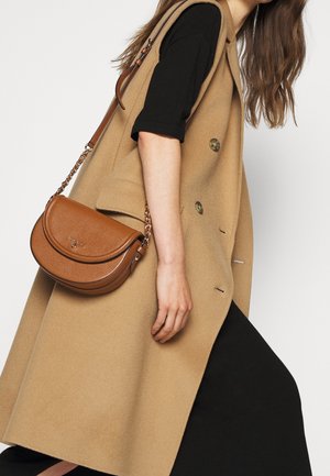 Femme portant une robe noire et un manteau sans manches à double boutonnage camel, portant un sac bandoulière en cuir marron avec une chaîne.