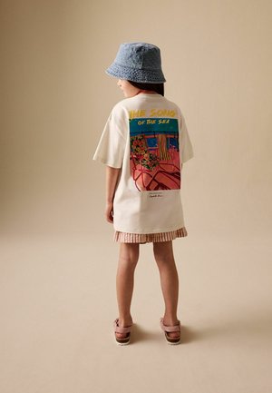 Laps, kes kannab beežikat üle suurusega T-särki, millel on värvikas "Merelaul" graafika, sinist ämbrimütsi, triibulisi lühse ja roosasid sandaale, seisab.