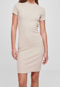 Robe beige ajustée à manches courtes avec un ourlet en frisottis sur les manches, le décolleté et l'ourlet, portée par une personne debout devant un fond uni.