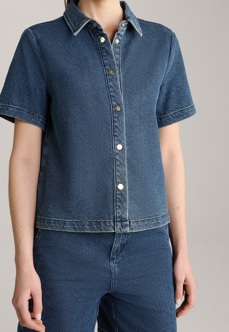 Personne portant une chemise en denim bleu à manches courtes avec col et un pantalon en denim bleu assorti, sur un fond uni.