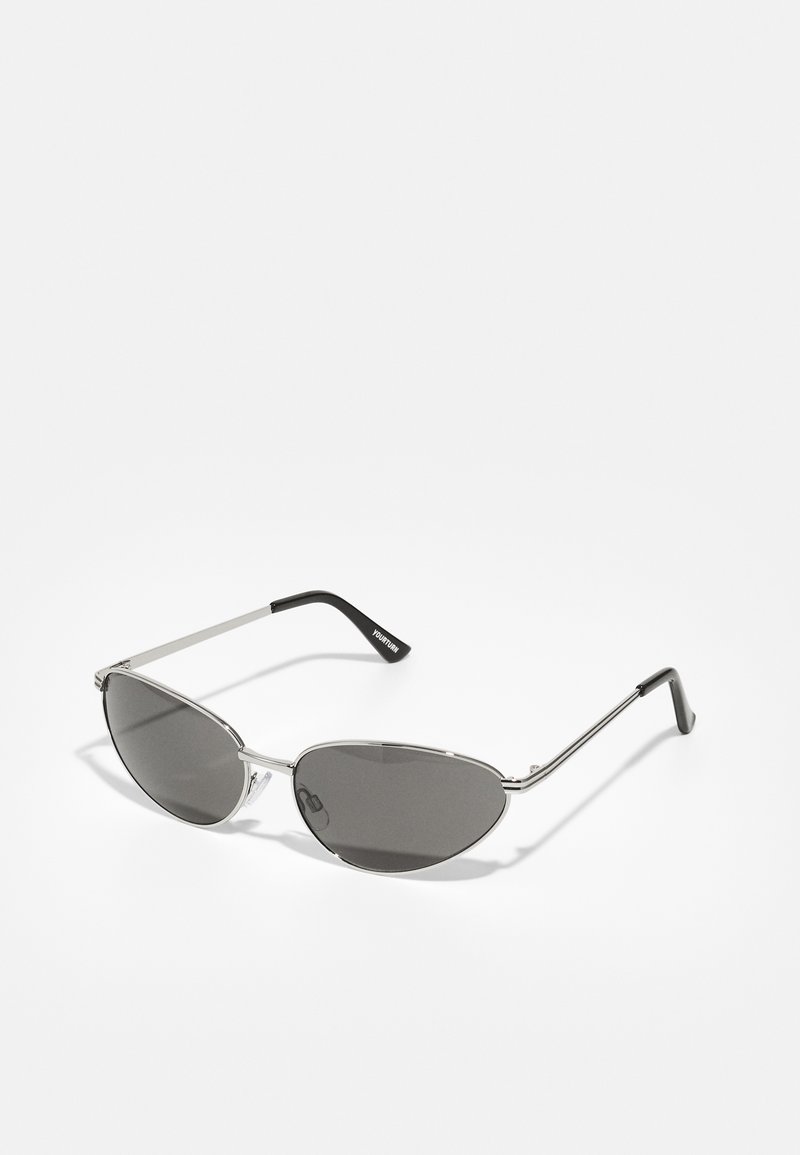 Lunettes de soleil avec une monture en métal argenté élégant, des verres teintés gris et un design aérodynamique avec des bords pointus et des branches fines.