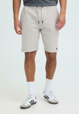 Homme portant un short gris clair à cordon de serrage, des chaussettes blanches montantes et des baskets blanches avec des rayures noires, debout sur un fond uni.