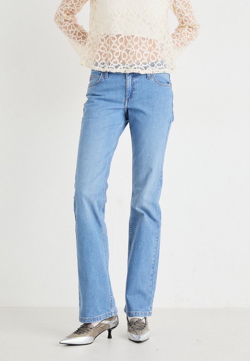 Jeans rectos de color azul claro con un diseño desgastado, estilo clásico de cinco bolsillos y detalles de costura sutiles, combinados con zapatos de tacón plateados.