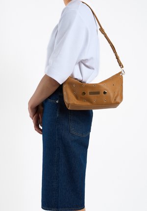Sac à épaule en cuir tan avec des rivets et des accents en métal. Forme semi-ovale, doté d'une fermeture éclair et d'une sangle ajustable. Porté avec une jupe en denim foncé.