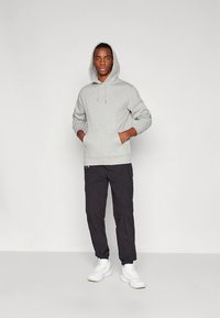 Felpa grigia con cappuccio e polsini a coste, abbinata a pantaloni neri aderenti e sneakers bianche, su sfondo semplice.