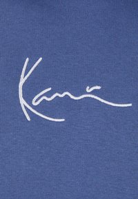 Tessuto blu con testo ricamato bianco che recita "Kari" in uno stile di scrittura fluido, con una texture liscia e un design pulito.