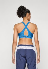 Sujetador deportivo azul con un diseño de espalda cruzada, que presenta un patrón texturizado y una banda elástica ancha etiquetada como "UNDER ARMOUR". Combinado con pantalones deportivos de color azul marino.