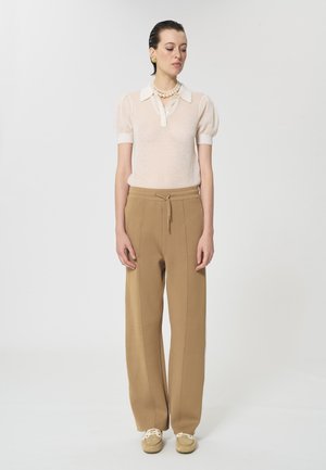 Femme portant un polo beige à manches courtes et transparentes, pantalon large beige avec cordon de serrage, chaussures beiges, collier de perles et boucles d'oreilles en or, debout.