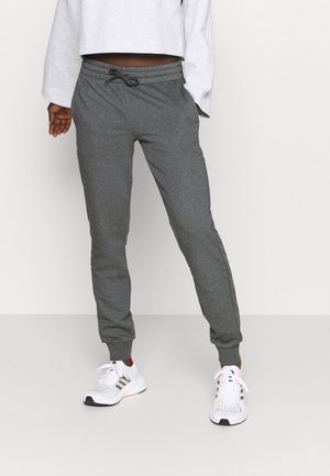 Person iført mørkegrå joggingbukser med snor, hvide sportssneakers og en cropped hvid langærmet top mod en ensfarvet baggrund.