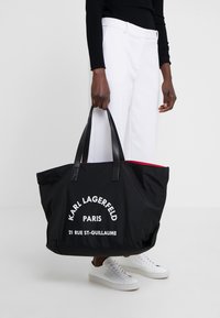 Personne portant un pantalon blanc et des baskets blanches tenant un grand sac fourre-tout noir avec le texte "Karl Lagerfeld Paris" en blanc.