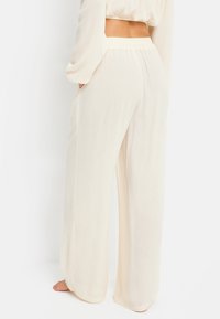 Woman wearing high-waisted, loose-fitting, cream-colored pants with an elastic waistband, standing barefoot against a white background.