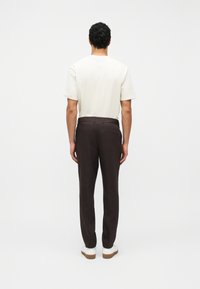 Homme debout de dos, portant une chemise blanche à manches courtes, un pantalon marron foncé avec une taille froncée, et des baskets blanches avec des semelles en caoutchouc.