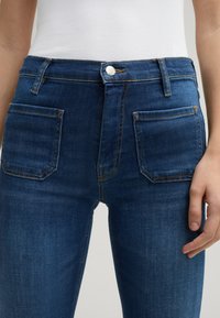 Niebieskie dżinsy z wysokim stanem, z dwoma przednimi kieszeniami i kontrastowymi pomarańczowymi szwami. Materiał ma gładką teksturę.