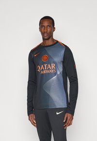 Uomo che indossa una maglia da calcio a maniche lunghe nera e grigia scura con accenti arancioni e il logo "Qatar Airways", abbinata a pantaloni Nike neri.