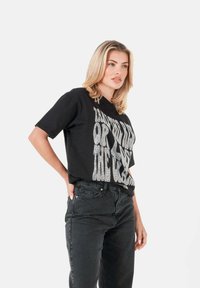 Camiseta negra de manga corta con texto gráfico en blanco llamativo. Combinada con pantalones vaqueros negros de cintura alta, que destacan un ajuste relajado y un estilo casual.