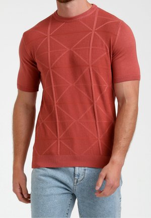 Homme portant un pull rouille à manches courtes avec un motif géométrique texturé et un jean bleu clair sur un fond uni.