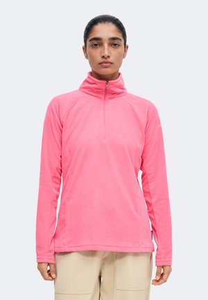 Kvinde står fremadvendt iført en knaldrød kvart-lynlås fleece top og beige bukser mod en ensartet lys baggrund.
