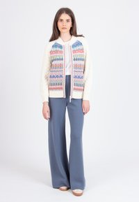 Cárdigan de punto con patrones geométricos multicolores, mangas color crema y cremallera completa. Combinado con pantalones de pierna ancha azules de talle alto.