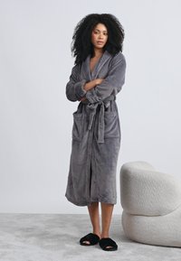 Pilka fliuso chalatas su šalkine apykakle, priekine raišteliu ir dviem priekinėmis kišenėmis. Modelis dėvi juodus naminius batus ant tekstūruotos šviesiai pilkos paviršiaus.