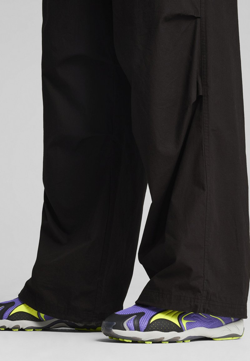 Black wide-leg pants with a smooth texture, paired with purple and yellow athletic shoes featuring a mesh upper and rugged sole.