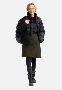Schwarze Steppjacke, olivgrüner Rock, schwarze Leggings und schwarze Schneestiefel. Strukturierte Oberflächen und kontrastreiche Farben schaffen einen geschichteten Look.