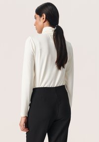Witte longsleeve top met een hoge nek en geraffineerde schouderdetails, gecombineerd met zwarte broeken, bekeken vanaf de achterkant. Glad textuur.