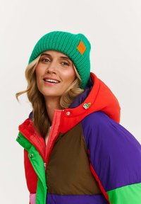 Jasne zelená rebrovaná čiapka s oranžovou nášivkou, spojená s viacfarebnou bundou s vankúšikmi s červenými, fialovými, zelenými a hnedými sekciami.