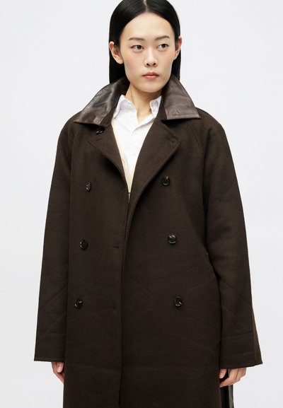 Γυναίκα με ίσια σκούρα μαλλιά φοράει καφέ διπλόχρωμο παλτό με δερμάτινο γιακά πάνω από λευκό πουκάμισο, κοιτάζοντας ελαφρώς δεξιά.