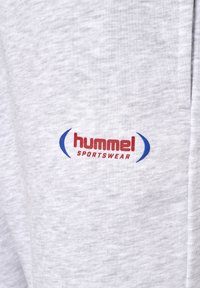 Graues Sweatshirtmaterial mit einem roten und blauen "hummel sportswear" Logo, das aus glattem, leicht strukturiertem Stoff besteht und ein typisches sportliches Design aufweist.
