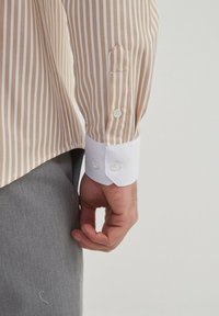 Chemise rayée beige et blanche avec une manchette blanche, comportant des boutons au poignet et une texture lisse. Un pantalon gris visible en arrière-plan.