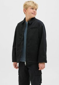 Blond pojke som bär en delvis zippad svart jacka över en blå skjorta och svarta cargobyxor, som tittar åt sin högra sida mot en enkel bakgrund.