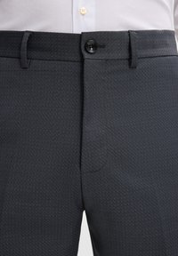 Ciemnoszare dopasowane spodnie z zapięciem na guziki i szlufkami, noszone z białą koszulą elegancką włożoną w spodnie.