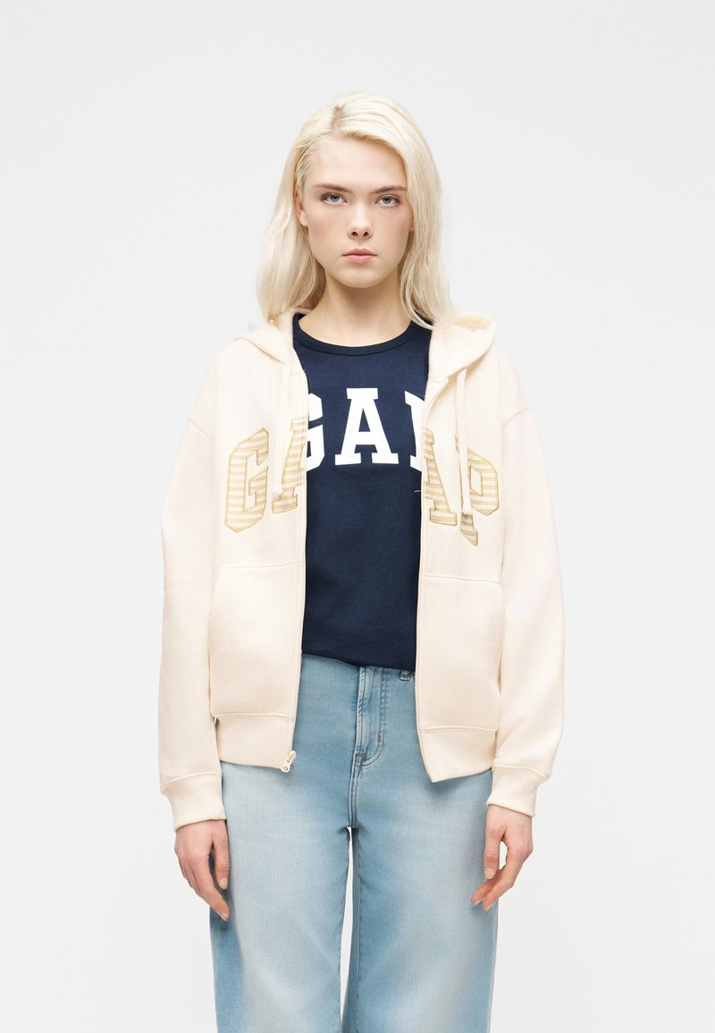 Νεαρή γυναίκα με ίσια ξανθά μαλλιά φοράει ανοιχτό μπεζ φούτερ με φερμουάρ πάνω από μπλε σκούρο t-shirt GAP και ανοιχτό γαλάζιο τζιν, σε ουδέτερο φόντο.