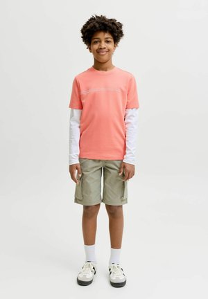 Garçon debout et souriant, portant une chemise corail à manches courtes sur un sous-pull blanc à manches longues, un short cargo kaki, des chaussettes blanches et des baskets noires et blanches.