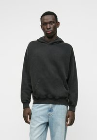 Hoodie noir en tissu doux, avec une poche frontale et un capuchon à cordon. Associé à un jean déchiré bleu clair.