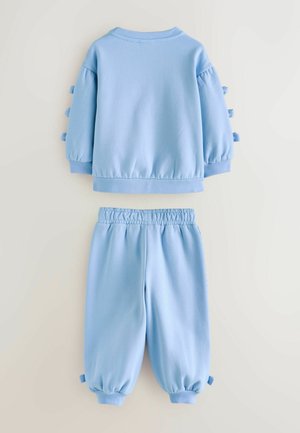 Světle modrá mikina s nabíranými rukávy a mašlí, doplněná o odpovídající kalhoty s elastickým pasem a manžetami na kotnících, vyrobené z měkké tkaniny.