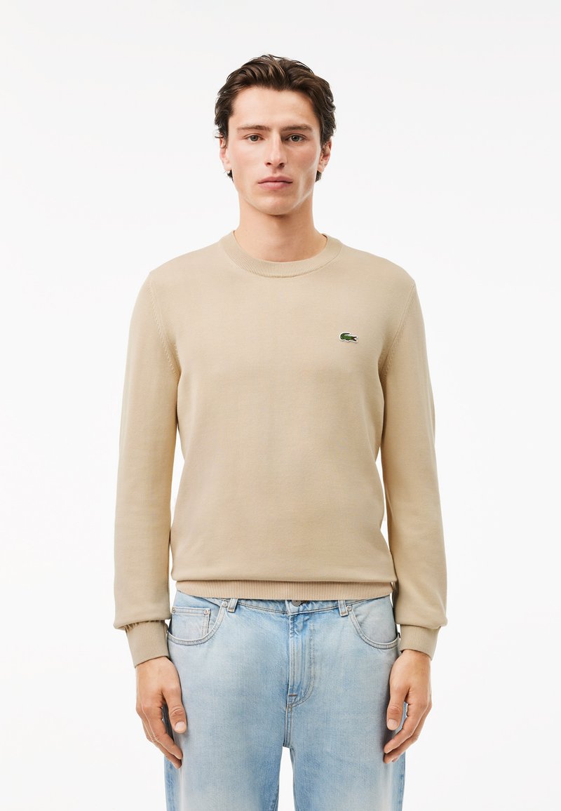 Beige gestrickter Pullover mit Rundhalsausschnitt, langen Ärmeln und gerippten Bündchen. Kleines grünes Krokodil-Logo auf der linken Brust, getragen zu hellblauen Jeans.