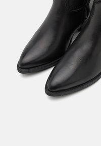 Botas de cuero negro hasta el tobillo con punteras puntiagudas, textura suave y un detalle de costura mínima a lo largo de los bordes. Suelas de goma negra planas.