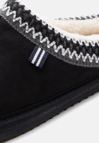 Crna papuča s mekim vunenim podstavom, sivi i bijeli zigzag šav s rubom i crna tkanina za povlačenje s bijelim prugama.