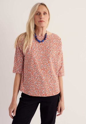 Frau mit langen blonden Haaren trägt eine rote Bluse mit Blumenmuster, eine blaue Perlenkette und eine schwarze Hose und steht vor einem schlichten, hellen Hintergrund.