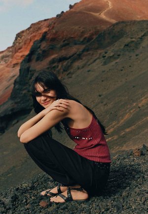 Jeune femme aux cheveux noirs portant un top rouge sans manches et un pantalon noir, assise sur un sol rocheux avec en arrière-plan une montagne rouge et noire.