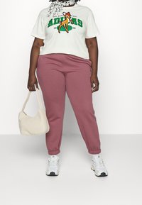 Valkoinen t-paita, jossa on vihreä ja oranssi Adidas-kuvio, yhdistettynä vaaleanpunaisiin verkkareihin, valkoisiin tennareihin ja pörröiseen vaaleankreemiin käsilaukkuun.