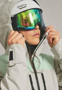 Ski goggles with reflective green lenses and white frame, worn with a light grey and mint green waterproof jacket, featuring a hood and zippers.