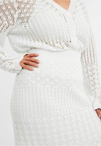 Weißer Strickcardigan mit komplexen Diamant- und Gittermustern, ausgestattet mit einer Knopfleiste und gerippten Bündchen, kombiniert mit einem passenden gestrickten Rock.