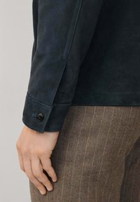 Marineblauwe suède jasmanchet met knoopdetail, in combinatie met bruine streepjesbroek. De gladde textuur en de getailleerde pasvorm zijn zichtbaar.